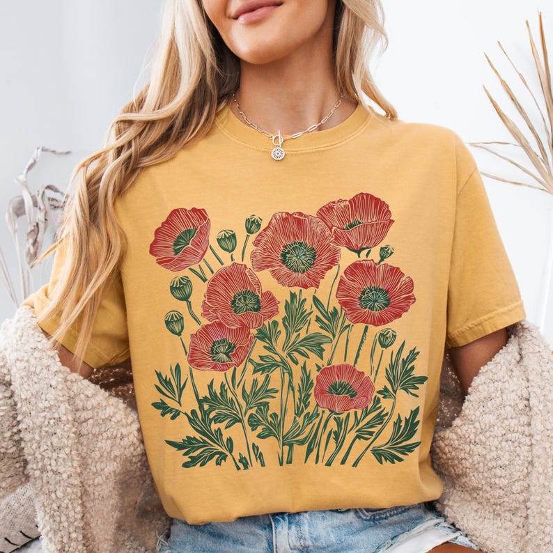 May include: Mustard yellow t-shirt featuring a red and green floral design. The graphic depicts several red poppies with green leaves and stems. The shirt has short sleeves and a crew neck.