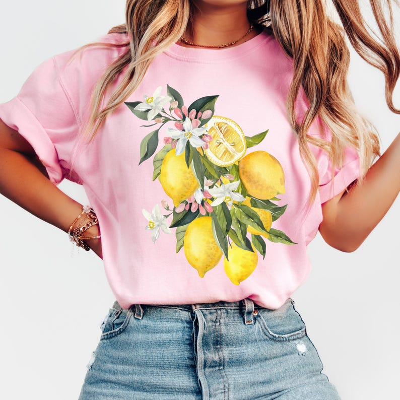 May include: A light pink t-shirt featuring a watercolor illustration of lemons, white blossoms, and green leaves. The lemons are yellow, and the blossoms have pink accents. The design is centered on the front of the shirt.