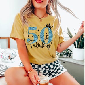 May include: A mustard yellow short-sleeve t-shirt featuring a graphic print with the text 50 & Fabulous in blue glitter, accented by a black crown icon and small star shapes. The shirt is paired with black and white checkered shorts.