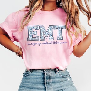 Puede incluir: Una camiseta rosa claro con las mangas dobladas. La camiseta presenta las letras "EMT" en un estampado floral azul, con "emergency medical technician" escrito debajo en una fuente cursiva. La modelo lleva vaqueros azules.