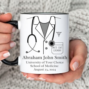 Pode incluir: Caneca de cerâmica branca com alça preta. A caneca apresenta uma ilustração em preto e branco de um jaleco médico com um estetoscópio, um bolso que diz "White Coat Ceremony", e o texto "Abraham John Smith, University of Your Choice, School of Medicine, August 23, 2024".