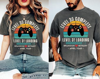 Op de afbeelding: Een grijs T-shirt met een retro videogamecontroller-afbeelding en de tekst "Level 30 voltooid" en "Level 31 wordt geladen". Het shirt bevat ook de tekst "Brandon Bethany - April Jaar -"