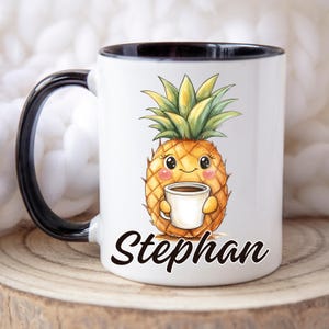 Op de afbeelding: Witte keramische mok met een zwart handvat en rand. De mok heeft een cartoon ananas die een koffiekopje vasthoudt, met de naam "Stephan" in zwart schrift eronder. De ananas heeft een lachend gezicht en roze wangen.