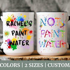 Puede incluir: Dos tazas de cerámica blanca con asas y bordes negros. Una taza dice "Rachel's Paint Water" con salpicaduras de pintura coloridas. La otra taza dice "Not Paint Water" con salpicaduras de pintura coloridas.