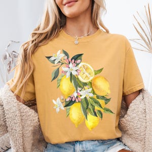 May include: Mustard yellow t-shirt featuring a watercolor illustration of lemons, white blossoms, and green leaves. The design includes whole and sliced lemons. The shirt has short sleeves and a crew neck.