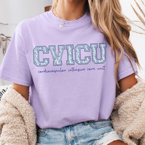 May include: A light purple t-shirt with the text "CVICU" in a blue floral design. Below the text is the phrase "cardiovascular intensive care unit" in a matching blue script. The shirt is short-sleeved.