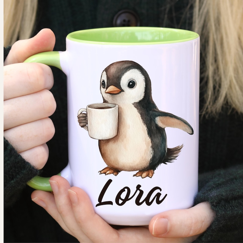 May include: White ceramic mug with a green interior and handle. The mug features a watercolor illustration of a penguin holding a coffee cup. The name "Lora" is written in black script below the penguin.