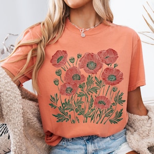 May include: A coral-colored t-shirt featuring a detailed illustration of red poppies with green leaves and stems. The floral design is centered on the shirt, creating a visually appealing botanical theme. The shirt has a classic crew neck and short sleeves.