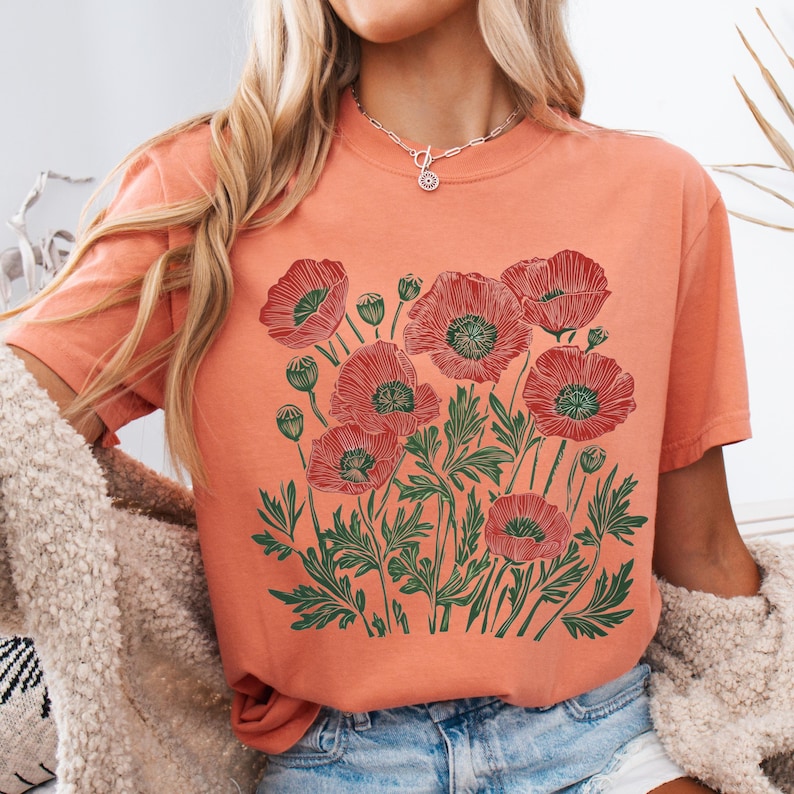 May include: A coral-colored t-shirt featuring a detailed illustration of red poppies with green leaves and stems. The floral design is centered on the shirt, creating a visually appealing botanical theme. The shirt has a classic crew neck and short sleeves.