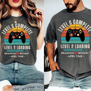 May include: A couple's matching t-shirts with a retro video game controller graphic. The shirts are gray with a faded look and the text "Level 8 Complete" and "Level 9 Loading".