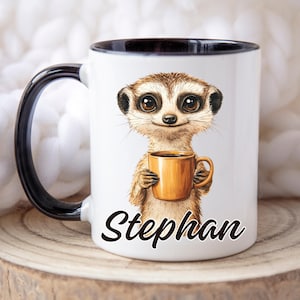May include: White ceramic mug with a black interior and handle. The mug displays a cartoon meerkat holding a gold coffee cup. The name "Stephan" is printed in black script on the front of the mug.