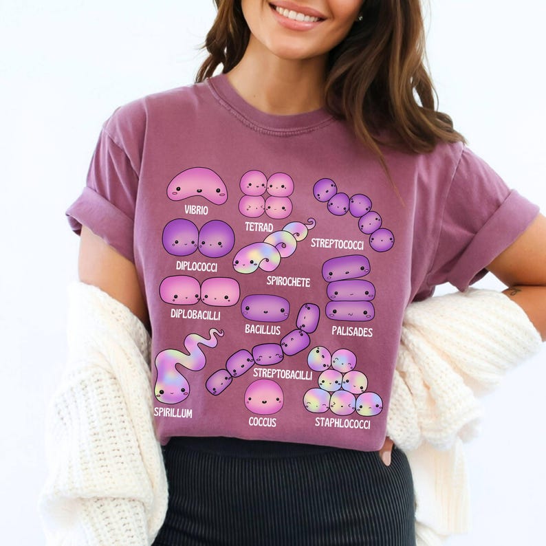 May include: A mauve t-shirt with a graphic of various bacteria, each depicted with a cute, cartoon-like design. The names of the bacteria, including Vibrio and Bacillus, are printed below each illustration.