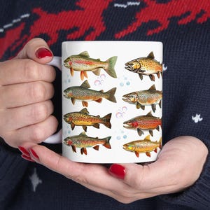 May include: White ceramic coffee mug featuring colorful illustrations of various fish species. The fish are depicted in shades of orange, yellow, brown, and green, with bubbles scattered around them. A unique gift for fishing enthusiasts.