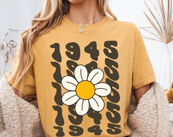 Può includere: T-shirt giallo senape con l'anno "1945" in un font retrò. Una grande margherita con centro giallo e petali bianchi è centrata sulla maglietta. La parola "SASSY" è stampata verticalmente sul lato destro.