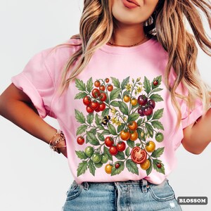 Op de afbeelding: Lichtroze T-shirt met een kleurrijke illustratie van een tomatenplant met rode, oranje, gele en groene tomaten, groene bladeren, een bij en lieveheersbeestjes. Het woord "BLOSSOM" staat in een witte doos rechtsonder.