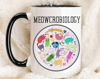Puede incluir: Taza de café de cerámica blanca con borde y asa negros. La taza presenta una ilustración colorida de gatos y el texto "MEOWCROBIOLOGY".