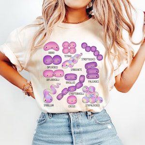 May include: A cream-coloured t-shirt displaying a vibrant illustration of different bacteria, such as Vibrio and Spirochete. Each bacterium is designed in a cute, cartoon style and labelled with its name in a playful font.