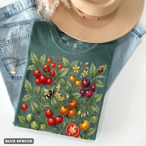 Op de afbeelding: Een tealkleurig t-shirt met een levendig tomatenplantontwerp met rode, oranje en groene tomaten, gele bloemen, een hommel en een lieveheersbeestje. Het shirt wordt gecombineerd met lichtblauwe jeans en een beige hoed. De tekst "BLUE SPRUCE" staat onderaan.