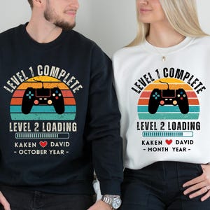 May include: Two black and white sweatshirts with a retro video game controller graphic. The text on the shirts reads "Level 1 Complete" and "Level 2 Loading".