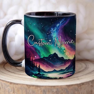 May include: A black and white ceramic mug featuring a colorful landscape design with mountains, trees, and a lake under a starry sky. The mug has a black handle and rim, and the text "Custom Name" is written on the front.