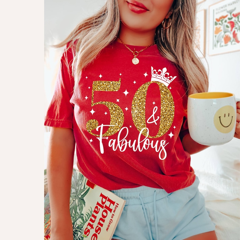 May include: A red short-sleeved t-shirt featuring a gold glitter graphic of the number 50, an ampersand, and the text "Fabulous" in white script, accented by a white crown icon and small star shapes printed on the front of the garment.