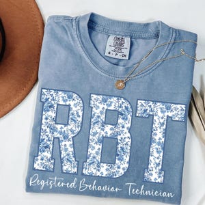 Könnte beinhalten: Ein blaues Comfort Colors T-Shirt mit den Buchstaben "RBT" in einem floralen Muster. Die Worte "Registered Behavior Technician" sind darunter gedruckt. Eine goldene Halskette liegt über dem Shirt.