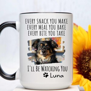 May include: White ceramic coffee mug with a black handle. The mug has a black rim and a black and white photo of a dog with the text "EVERY SNACK YOU MAKE EVERY MEAL YOU BAKE EVERY BITE YOU TAKE I'LL BE WATCHING YOU Luna" printed on it.