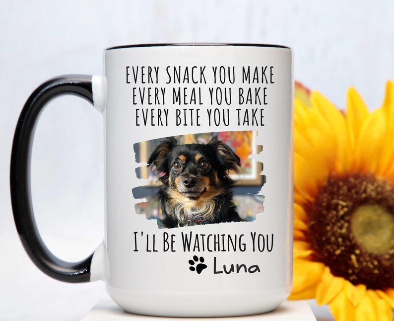 May include: White ceramic coffee mug with a black handle. The mug has a black rim and a black and white photo of a dog with the text "EVERY SNACK YOU MAKE EVERY MEAL YOU BAKE EVERY BITE YOU TAKE I'LL BE WATCHING YOU Luna" printed on it.