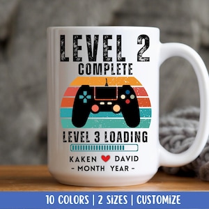May include: White ceramic mug with a handle, featuring the text "LEVEL 2 COMPLETE" above a retro-style controller graphic. Below, it reads "LEVEL 3 LOADING" with a progress bar, names, and a date. The mug is designed for couples.