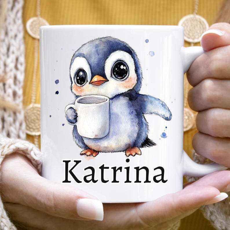 May include: White ceramic mug featuring a watercolor illustration of a cute penguin holding a coffee cup. The penguin is blue and gray with large eyes and a small orange beak. The name "Katrina" is printed in black below the penguin.