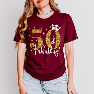 May include: A maroon short-sleeved t-shirt featuring the gold glitter text 50 & Fabulous with a white crown graphic above the zero. The design includes small white star accents scattered around the numbers on the front of the garment.