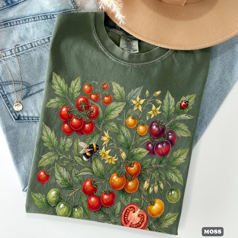 Op de afbeelding: Mosgroen T-shirt met een botanische illustratie van tomaten, bladeren en bloemen. Het ontwerp bevat rode, oranje en groene tomaten, een hommel en een lieveheersbeestje. Het shirt wordt gecombineerd met een blauwe jeans, een ketting en een beige hoed.