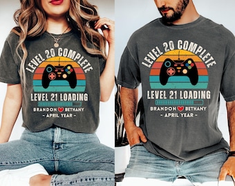 Op de afbeelding: Twee grijze T-shirts met een retro videogamecontroller-afbeelding en de tekst "Level 20 voltooid" en "Level 21 wordt geladen". De shirts bevatten ook de tekst "Brandon Bethany - April Jaar - "