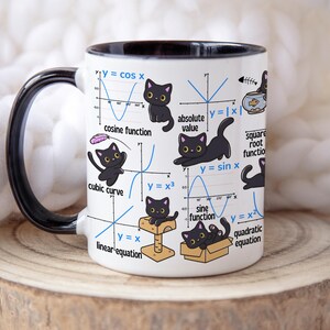 Puede incluir: Taza de cerámica blanca con asa y borde negros, con ilustraciones de gatos negros y gráficos matemáticos. La taza incluye ecuaciones como y=cos x e y=sin x. La taza se muestra sobre una superficie de madera.