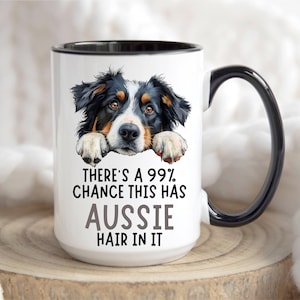 May include: A white ceramic mug with a black handle and rim featuring a colorful illustration of an Australian Shepherd dog resting its paws on the text. The mug displays the black text: There's a 99% chance this has Aussie hair in it.
