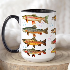 May include: White ceramic mug featuring colorful illustrations of various trout species. The mug has a black handle and rim, with bubbles scattered around the fish. The design includes rainbow trout, brown trout, and other fish, making it a great gift for fishing enthusiasts.
