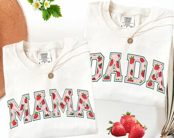 May include: Two off-white Comfort Colors t-shirts with the words "MAMA" and "DADA" in a strawberry pattern. Each shirt has a gold necklace. Fresh strawberries and white flowers are also in the image.