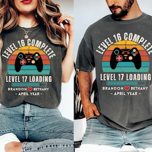 May include: Two gray t-shirts with a retro video game controller graphic and the text "Level 16 Complete" and "Level 17 Loading". The shirts also include the text "Brandon Bethany - April Year - "