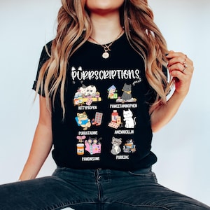 Könnte beinhalten: Schwarzes T-Shirt mit dem Text "Purrscriptions" und Illustrationen von Katzen mit verschiedenen Medikamenten. Das Shirt zeigt Cartoon-Katzen mit Namen wie Kittyprofen und Purrzac sowie Pillenflaschen.