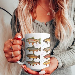 May include: White ceramic mug featuring colorful illustrations of various fish species. The fish designs include rainbow trout, brown trout, and other types, with bubbles around them. The mug is held by a person.