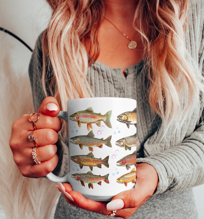 May include: White ceramic mug featuring colorful illustrations of various fish species. The fish designs include rainbow trout, brown trout, and other types, with bubbles around them. The mug is held by a person.