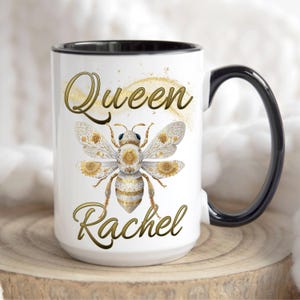 May include: White ceramic mug with a black handle and rim. The mug features a golden bee design with the words "Queen Rachel" in gold script. The bee has intricate details and is set against a white background.