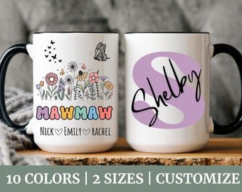 May include: Two white ceramic mugs with black handles. One mug features a floral design with the text "Mawmaw Nick Emily Rachel". The other mug has a purple swirl with the text "Shelby".