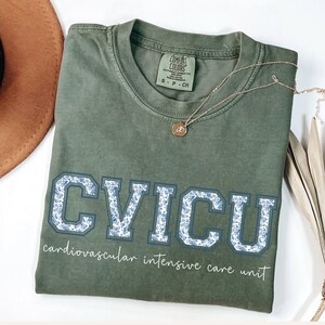May include: Olive green t-shirt with the letters "CVICU" in a blue floral design. The words "cardiovascular intensive care unit" are printed below the letters. A gold necklace and a brown hat are also visible.