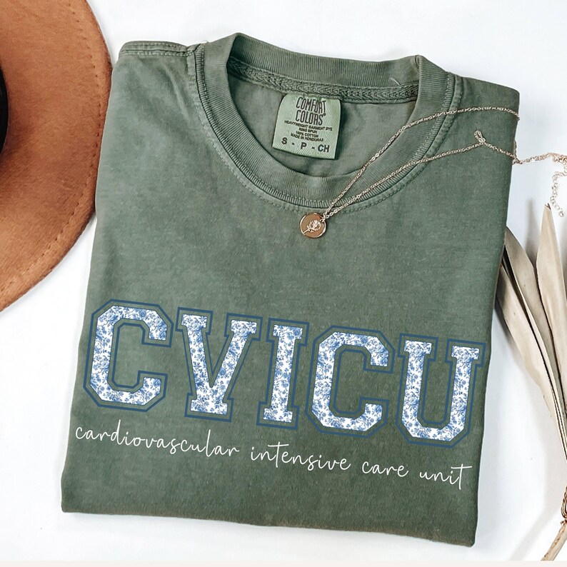 May include: Olive green t-shirt with the letters "CVICU" in a blue floral design. The words "cardiovascular intensive care unit" are printed below the letters. A gold necklace and a brown hat are also visible.