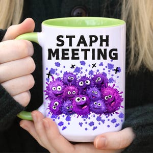 May include: White ceramic mug with a green interior and handle. The mug features the text "STAPH MEETING" in black, along with a cartoon illustration of purple, smiling bacteria. Purple confetti and black crosses are scattered around the design.