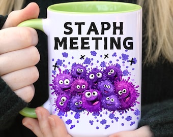 May include: White ceramic mug with a green interior and handle. The mug features the text "STAPH MEETING" in black, along with a cartoon illustration of purple, smiling bacteria. Purple confetti and black crosses are scattered around the design.