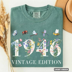 Op de afbeelding: Een saliegroen t-shirt met de witte tekst 1946 VINTAGE EDITION. Het ontwerp bevat grote witte cijfers gevuld met kleurrijke illustraties van wilde bloemen en een rij insecten, waaronder een vlinder, lieveheersbeestje, libel en bij.
