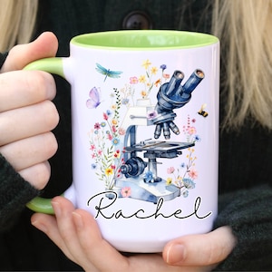 Op de afbeelding: Witte keramische mok met een groene rand. De mok heeft een aquarel-illustratie van een microscoop met bloemen en een libel. De tekst "Rachel" is in zwarte cursieve letters onder de afbeelding geschreven.