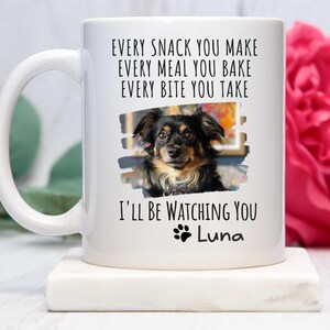 May include: White ceramic coffee mug with a black paw print and the text "EVERY SNACK YOU MAKE EVERY MEAL YOU BAKE EVERY BITE YOU TAKE I'LL BE WATCHING YOU Luna". A photo of a brown and black dog is in the center of the mug.